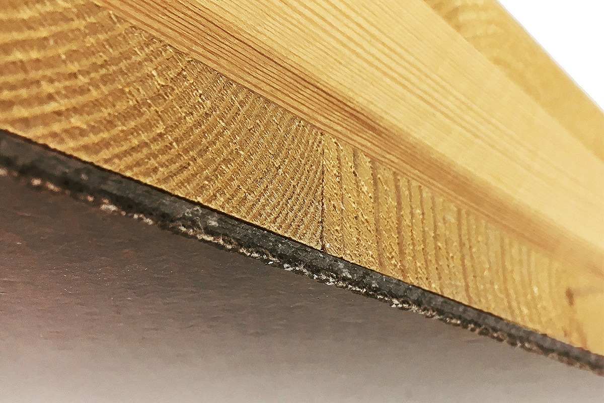 The photo shows the cut edge of a wooden panel whose underside is completely covered with a gray-brown material. 