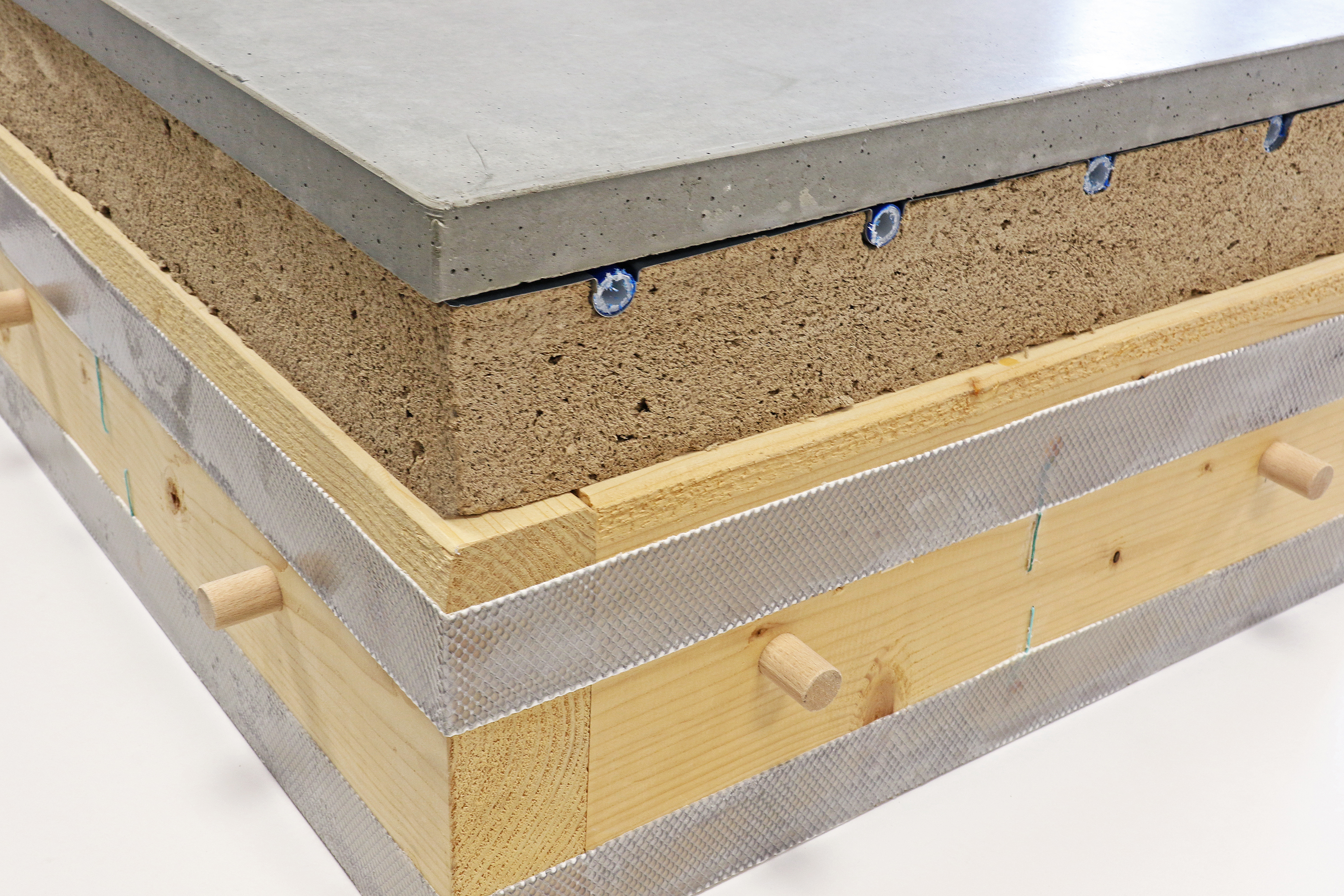 The photo shows a wooden box with a height of around 15 cm. Adhesive tape is attached to the side of the box and round wooden rods protrude as plug connections. On top of the box sits a 10 cm-high wood-foam board covered by a 4 cm-thick layer of concrete. Directly under the concrete layer, pipes are embedded in the wood foam.
