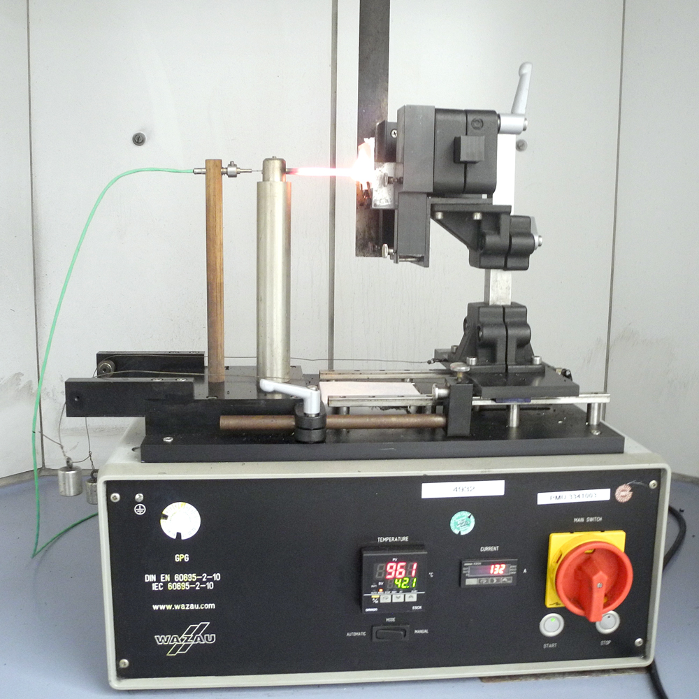 Versuchsapparatur, in die ein kleiner Prüfkörper aus Bio-Kunststoff eingespannt ist. Der anliegende Draht ist weiß glühend.