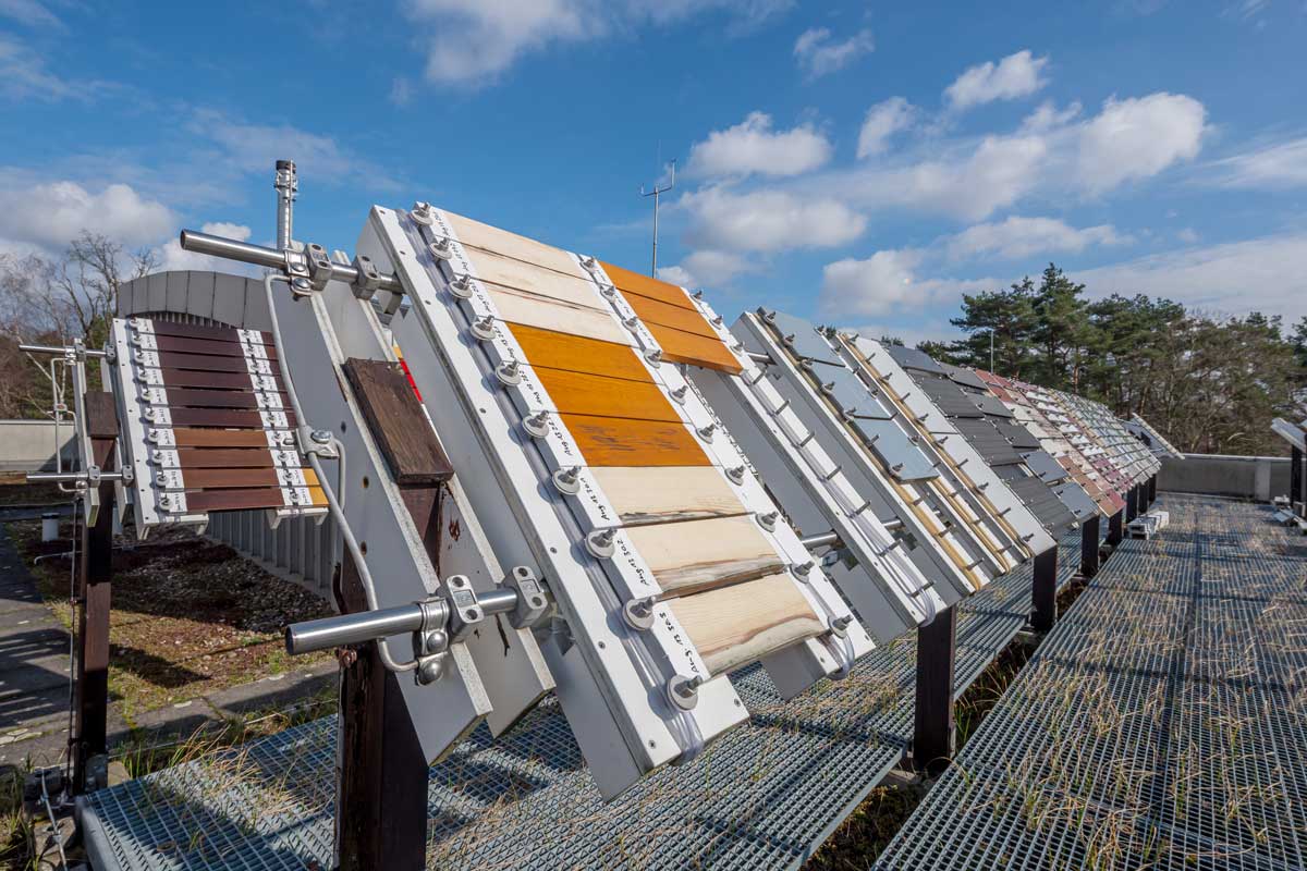 Das Foto zeigt ein Metallgestell unter freiem Himmel, das mit einer Vielzahl an verschiedenfarbig beschichteten Holzteilen bestückt ist, die schräg Richtung Himmel geneigt sind.