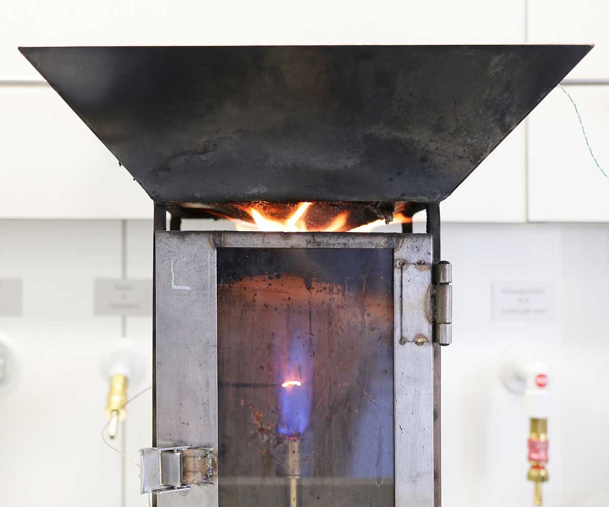 Das Foto zeigt einen metallischen Kasten mit Luftschlitzen und einer Glastür, durch die man eine Bunsenbrennerflamme im Inneren des Kastens sieht. An der Decke des Kastens befindet sich ein Sperrholz, welches mit dem neu entwickelten intumeszierenden Lack beschichtet wurde. Diese Beschichtung ist hier durch das Einwirken der Gasflamme aufgeschäumt, wodurch ein schwarz-braunes, schaumartiges Material entstanden ist, das sich hügelförmig nach unten wölbt und von Flammen umzüngelt wird.