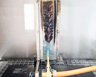 Das Foto zeigt eine Laborapparatur: Auf einer vertikal eingespannten Metallplatte ist eine Klebstofffolie aufgebracht, die mithilfe einer Gasbrenner-Flamme von schräg unten beflammt wird. 