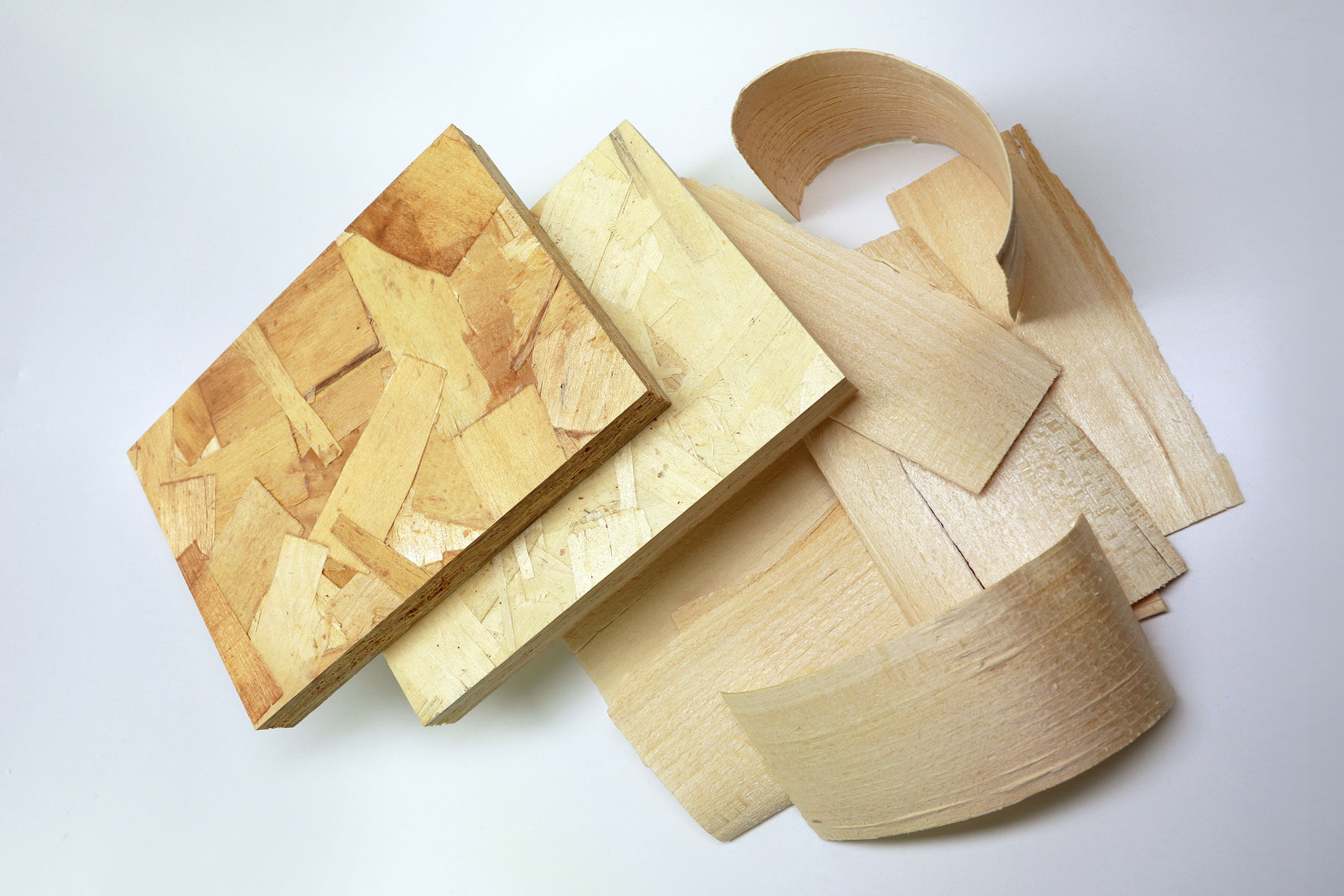 Das Foto zeigt zwei OSB-Plattenstücke aus hellem sowie etwas dunklerem Holz sowie einen Haufen Grobspäne (»Strands«) aus Laubhholz.