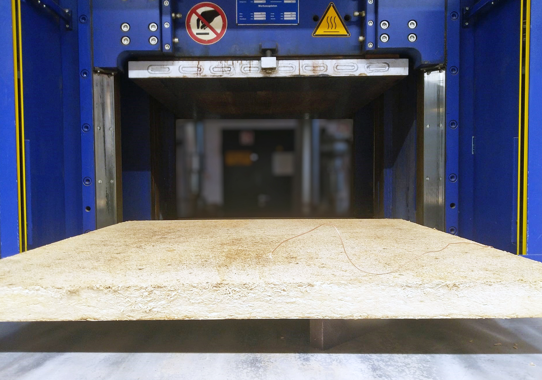 Pilzmyzel-Wärmedämmplatte aus Heißpresse Das Foto zeigt eine 1 x 1 Meter große und 9 cm starke Platte mit beige-hellbrauner Farbe, die auf einem Tisch liegt. Im Hintergrund ist eine Plattenpresse zu sehen.