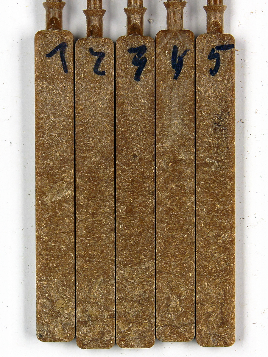 Das Foto zeigt fünf etwa 8 cm lange, flache Stäbe aus einem karamellbraunen Material mit feinkörniger Struktur (WPC). 