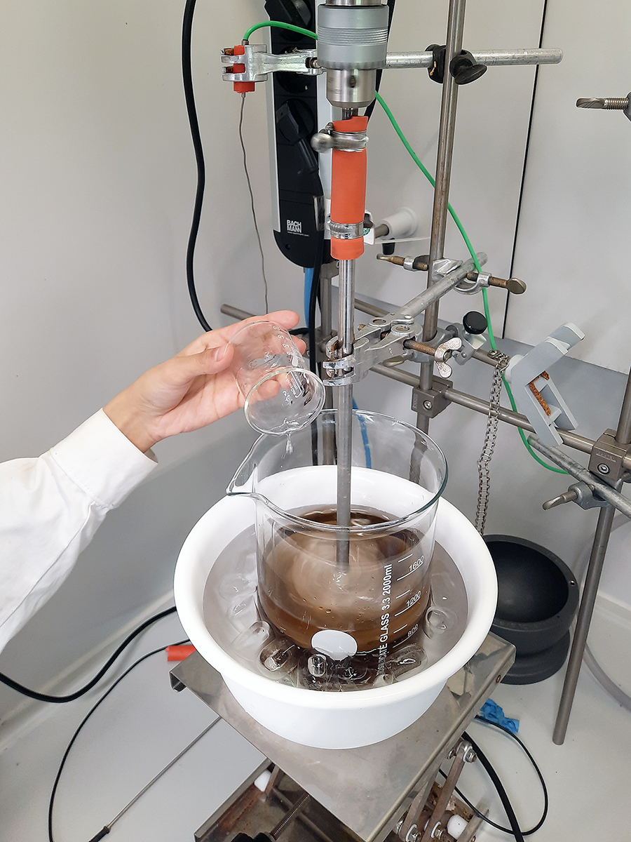 Synthese von biobasierten Ammonium-Phytate im Chemielabor (Projekt »ScaleAmp«) Das Foto zeigt ein Becherglas in einem Eiswürfelbad. Das Laborglas enthält eine dunkelbraune Flüssigkeit, in die gerade eine transparente Flüssigkeit gegossen wird. Ein elektrischer Rührstab verquirlt beide Flüssigkeiten miteinander. Man erkennt die beginnende Bildung eines Feststoffes, der durch die chemische Reaktion aus der Lösung ausfällt.