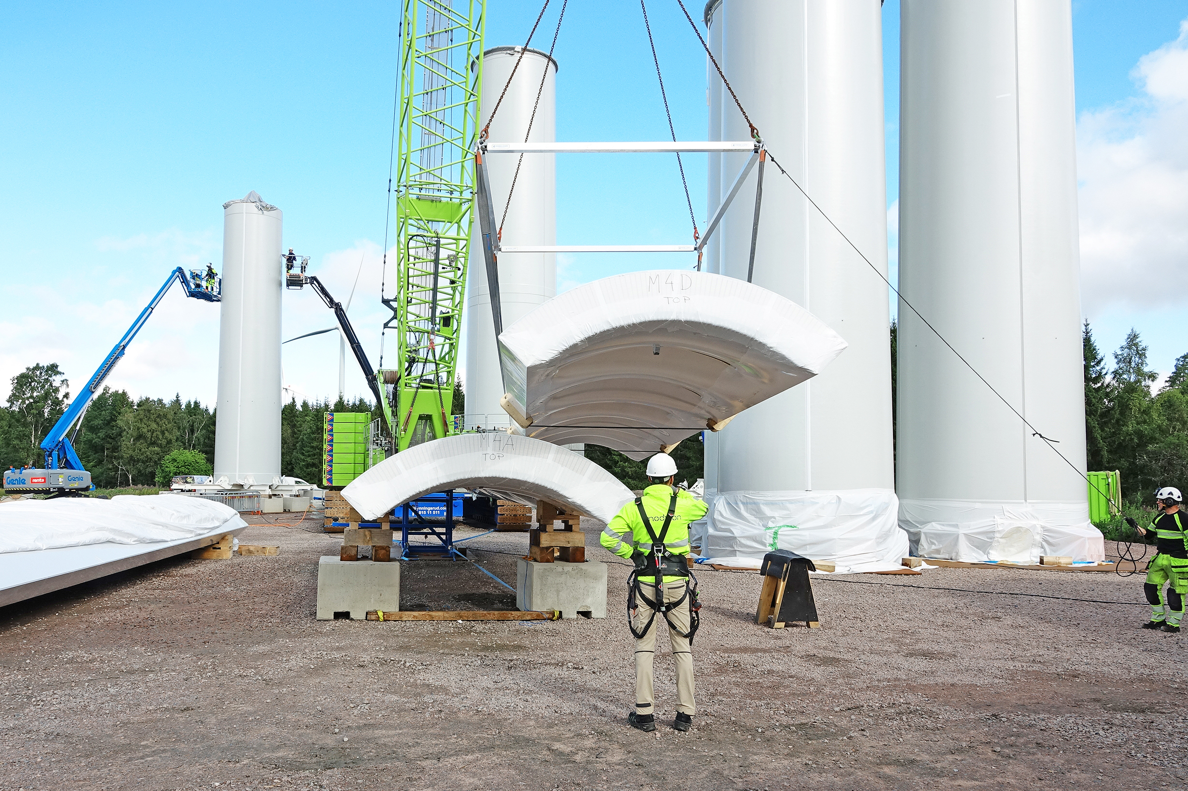 Bauteil-Anlieferung für einen Windkraft-Holzturm (Modvion) Das Foto zeigt eine Baustelle mit einem Bauarbeiter in der Mitte und einer Viertelschale, die von einem Kran bewegt wird.