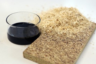 Das Foto zeigt ein Stück Spanplatte neben einem Laborglas mit einer zähen, schwarzbraunen Flüssigkeit sowie einem kleinen Haufen Holzspäne. 