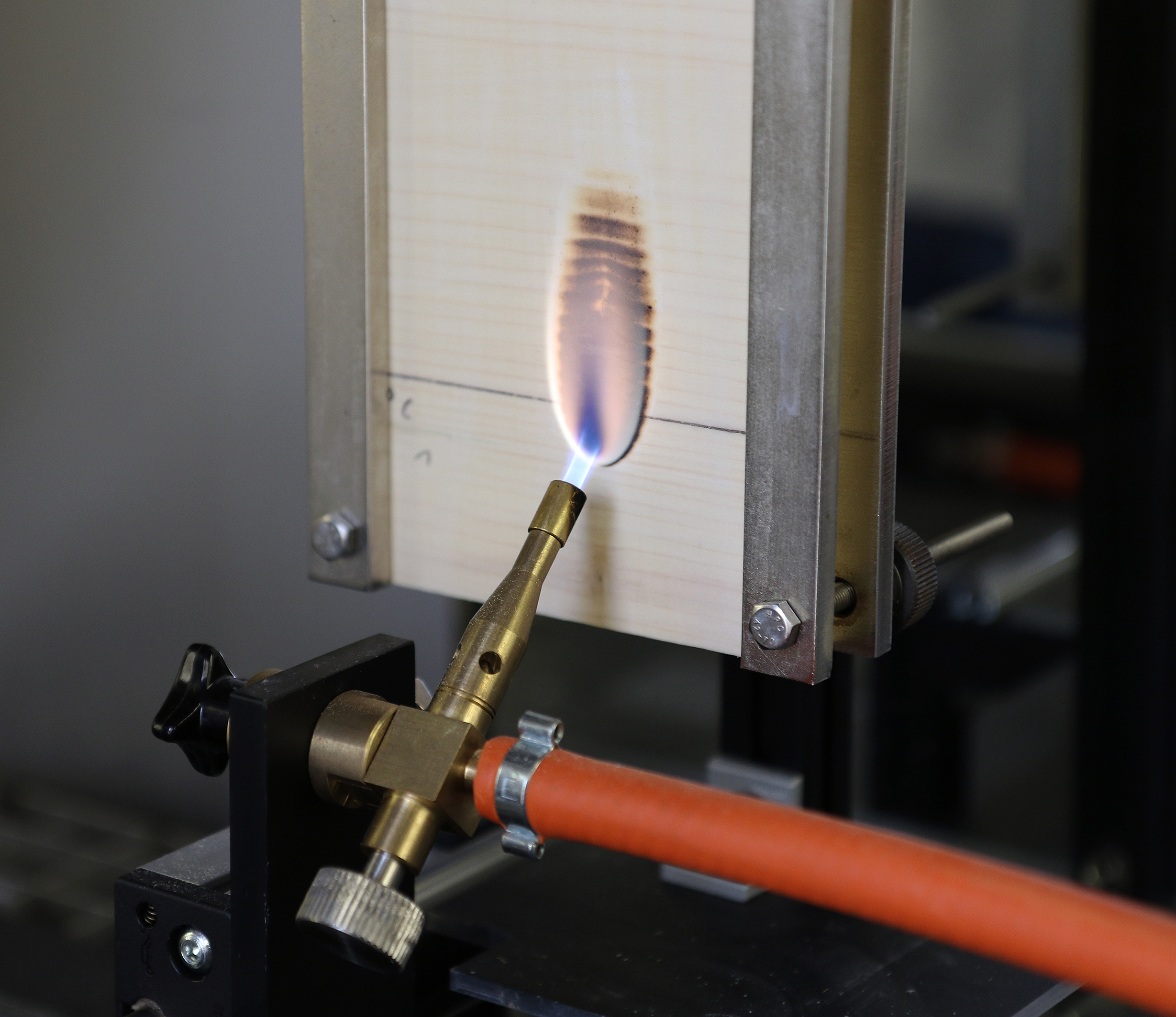 Beflammungstest einer beschichteten Holzprobe Das Foto zeigt eine metallische Apparatur, in die ein Stück Fichtensperrholz eingespannt ist. Das Holzstück wird mithilfe einer davor installierten Gasflamme direkt beflammt. Auf dem Holz bildet sich im Bereich der Beflammung ein ovaler dunkelbrauner Fleck (Verkohlung).
