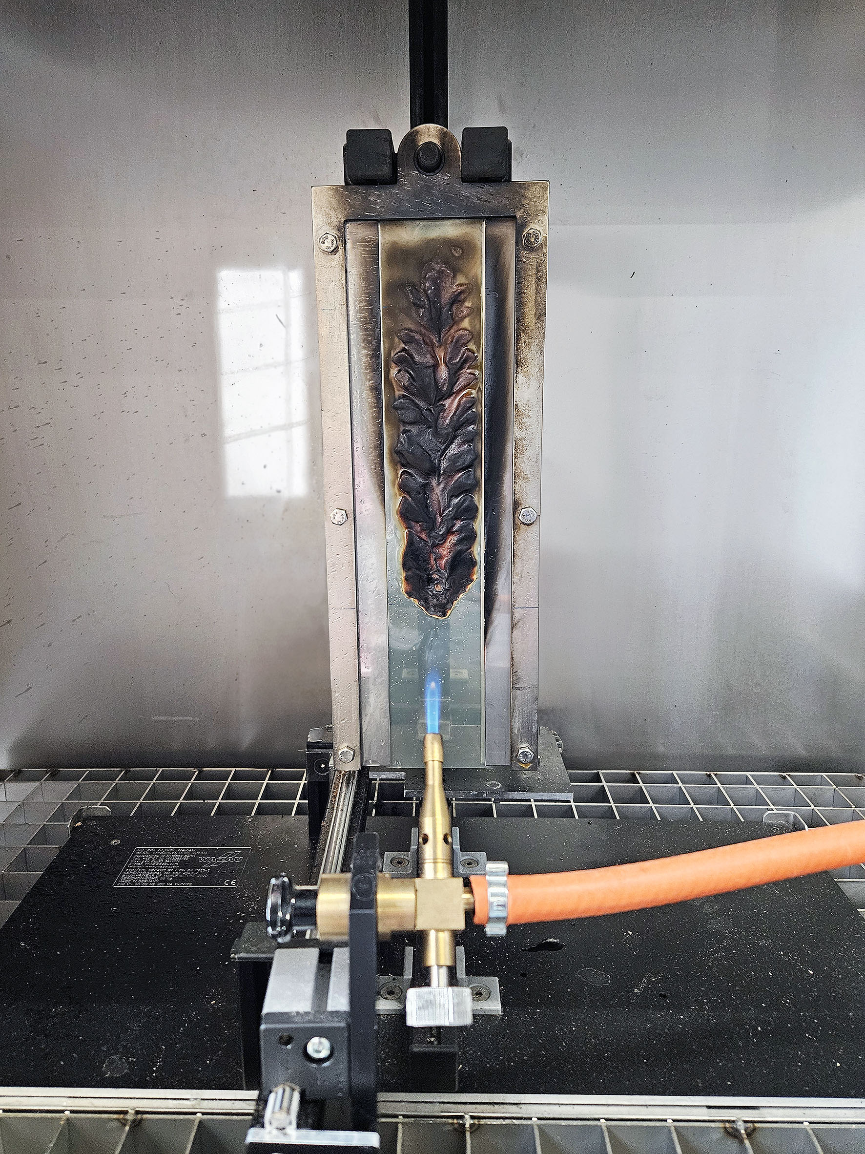 Modifizierter Einzelflammentest an einer duroplastischen Klebstofffolie (Projekt »FireSafe-CLT«) Das Foto zeigt eine Laborapparatur: Auf einer vertikal eingespannten Metallplatte ist eine Klebstofffolie aufgebracht, die mithilfe einer Gasbrenner-Flamme von schräg unten beflammt wird.
