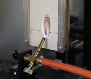 Das Foto zeigt eine metallische Apparatur, in die ein Stück Fichtensperrholz eingespannt ist. Das Holzstück wird mithilfe einer davor installierten Gasflamme direkt beflammt. Auf dem Holz bildet sich im Bereich der Beflammung ein ovaler dunkelbrauner Fleck (Verkohlung).