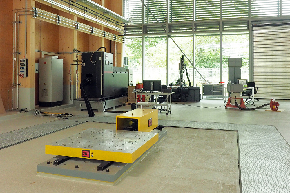 Das Foto zeigt eine Halle von innen. Im Hallenboden ist ein rechteckiges Fundament eingelassen, auf dem der Shaking Table steht. Der Shaking Table besteht aus einem 1,5 x 1,5 Meter großen, massiven Platte und einem daran angeschlossenen Motorblock.