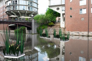 Das Bild ist eine Kombination aus einem Foto (Hintergrund) und grafischen Visualisierungen. Das Foto zeigt einen Wasserkanal in Hamburg (Fleet). Auf dem Wasser schwimmen mehrere kleine, bepflanzte Inseln (Computergrafik).
