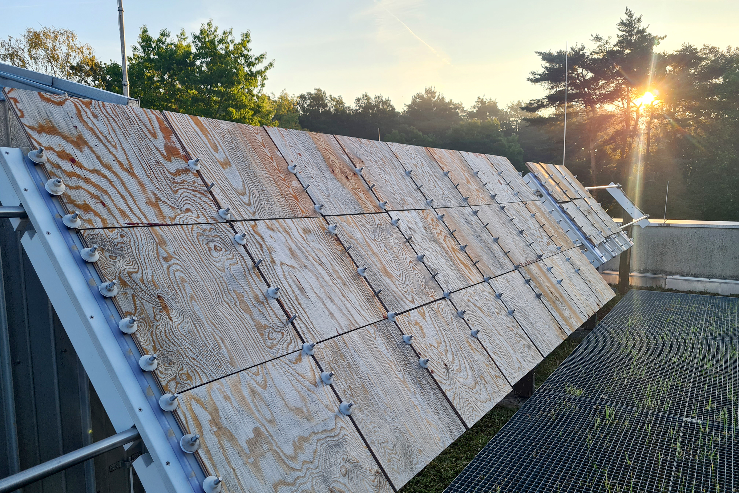 Das Foto zeigt ein Metallgestell unter freiem Himmel, das mit einer Vielzahl an Holzplattenstücken aus Sperrholz bestückt ist, die schräg Richtung Himmel geneigt sind.