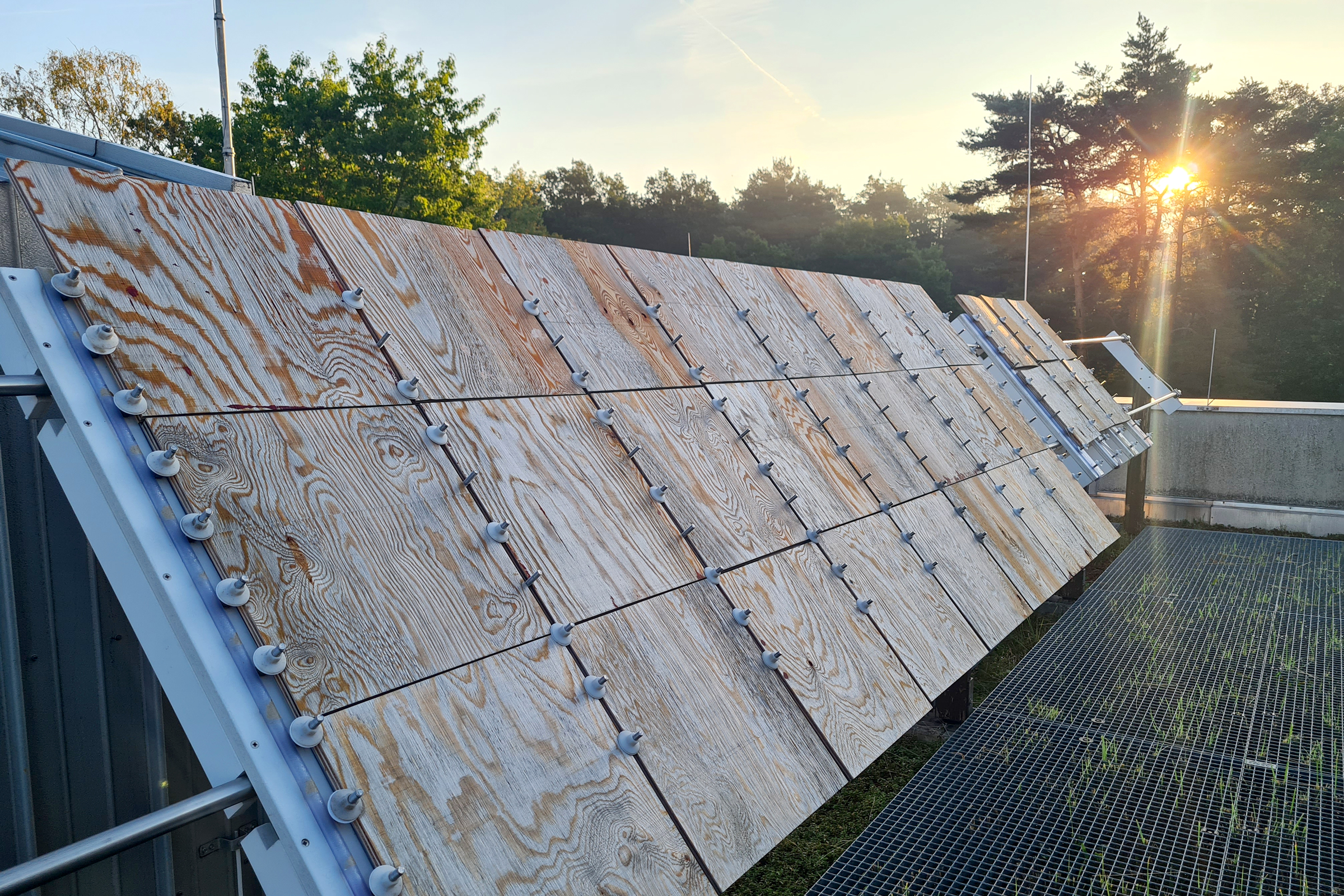 Das Foto zeigt ein Metallgestell unter freiem Himmel, das mit einer Vielzahl an Holzplattenstücken aus Sperrholz bestückt ist, die schräg Richtung Himmel geneigt sind.