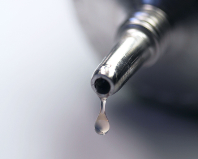 The tip of a silver-colored tube of adhesive out of which a drop of transparent adhesive drips.