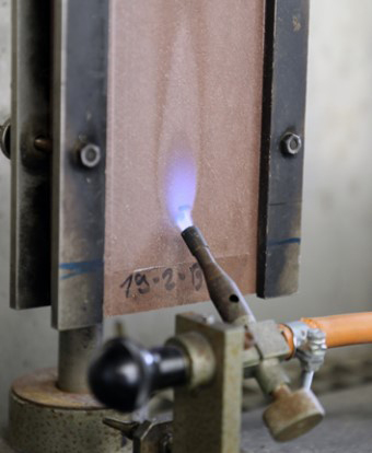 Testing apparatus with a clamped piece of natural material which is being flame-treated using a Bunsen burner.