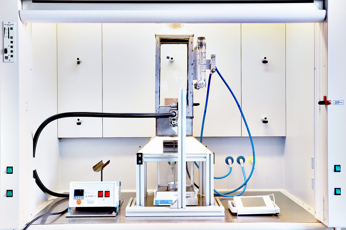 The photo shows a technical apparatus with a chamber containing a wooden test specimen measuring approximately 10 x 10 cm.