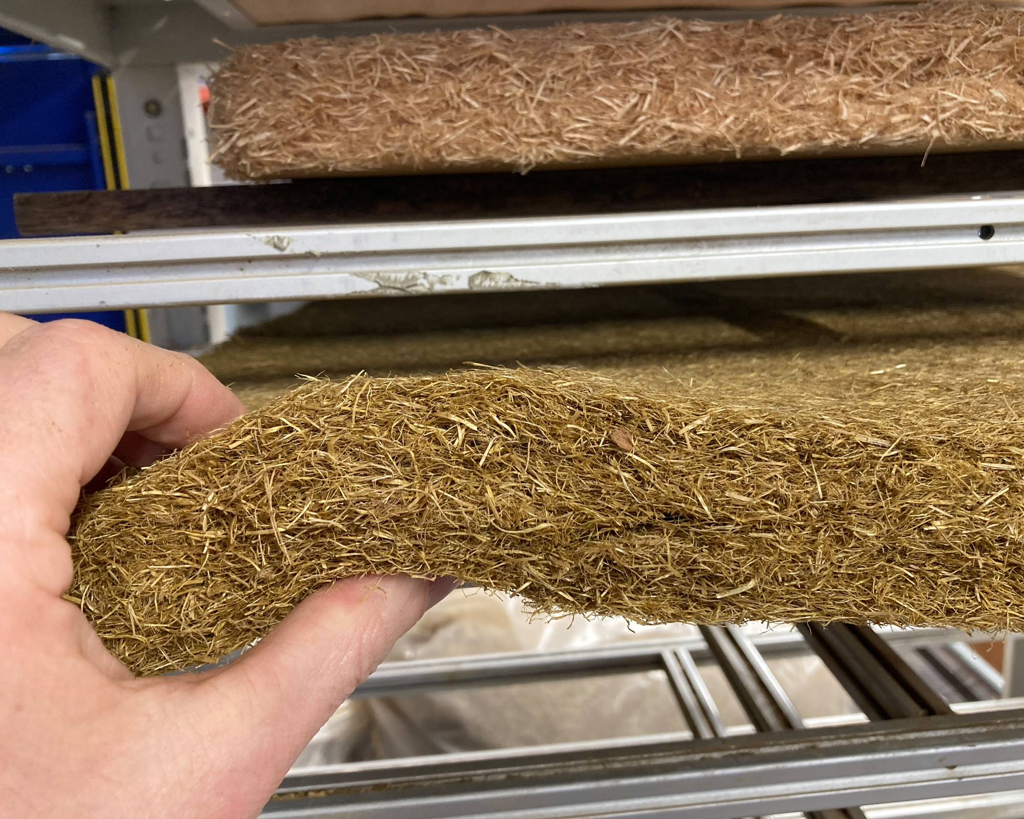 Flexible insulation mats made from wheat straw The photo shows a hand which is holding and slightly bending an approximately 5 cm thick flexible insulation mat made from wheat straw.