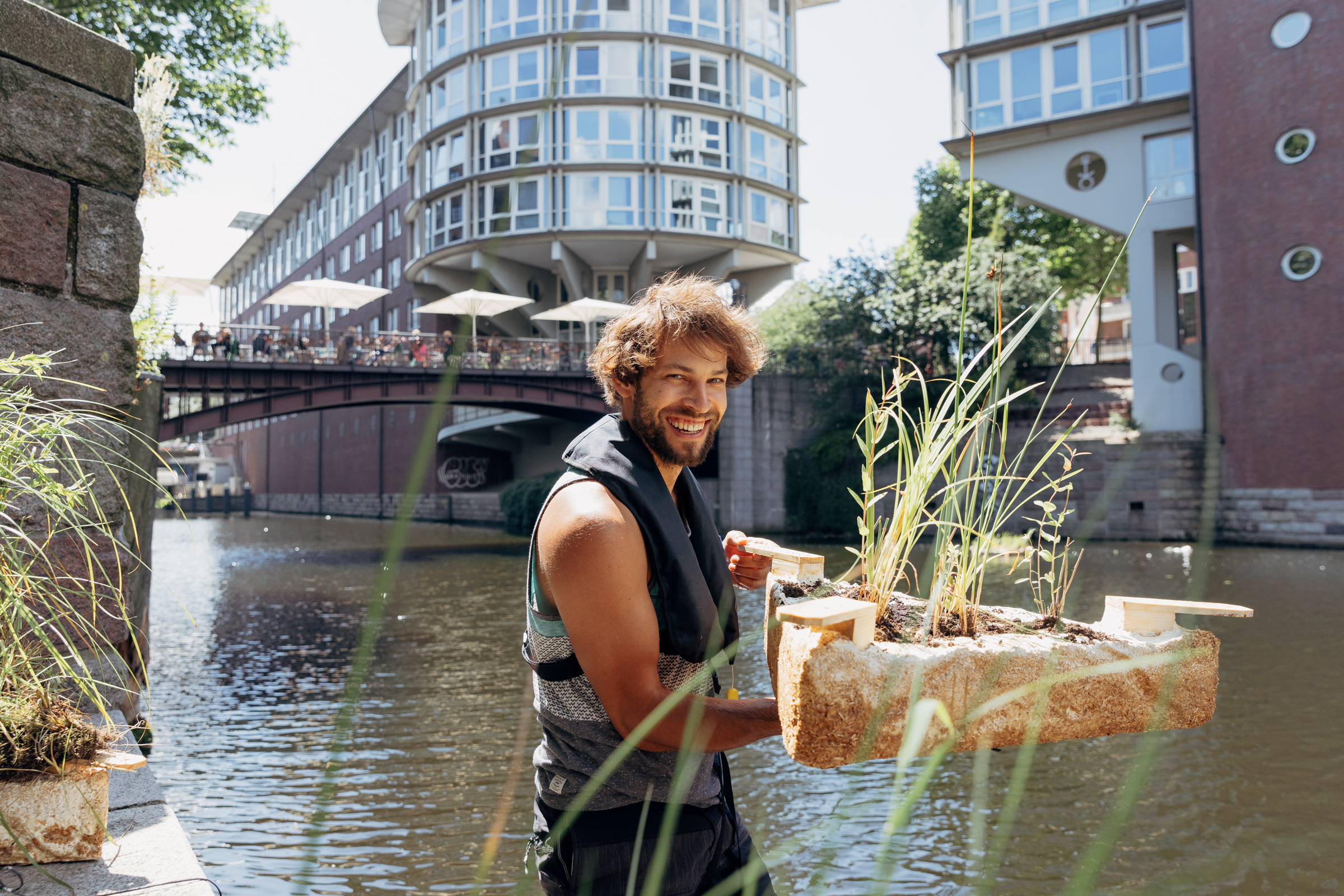 Project “Grünes Wasser” (Green Water): Launch in the Bleichenfleet in Hamburg The photo shows a researcher holding a triangular floating island made from mycelium material, in which plants are growing. At the corners, connecting elements made from wood can be seen.