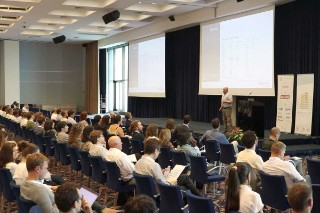 The picture shows Professor Bohumil Kasal during his keynote lecture. He is standing on the stage of a conference hall. Around 50 people are sitting in the audience.