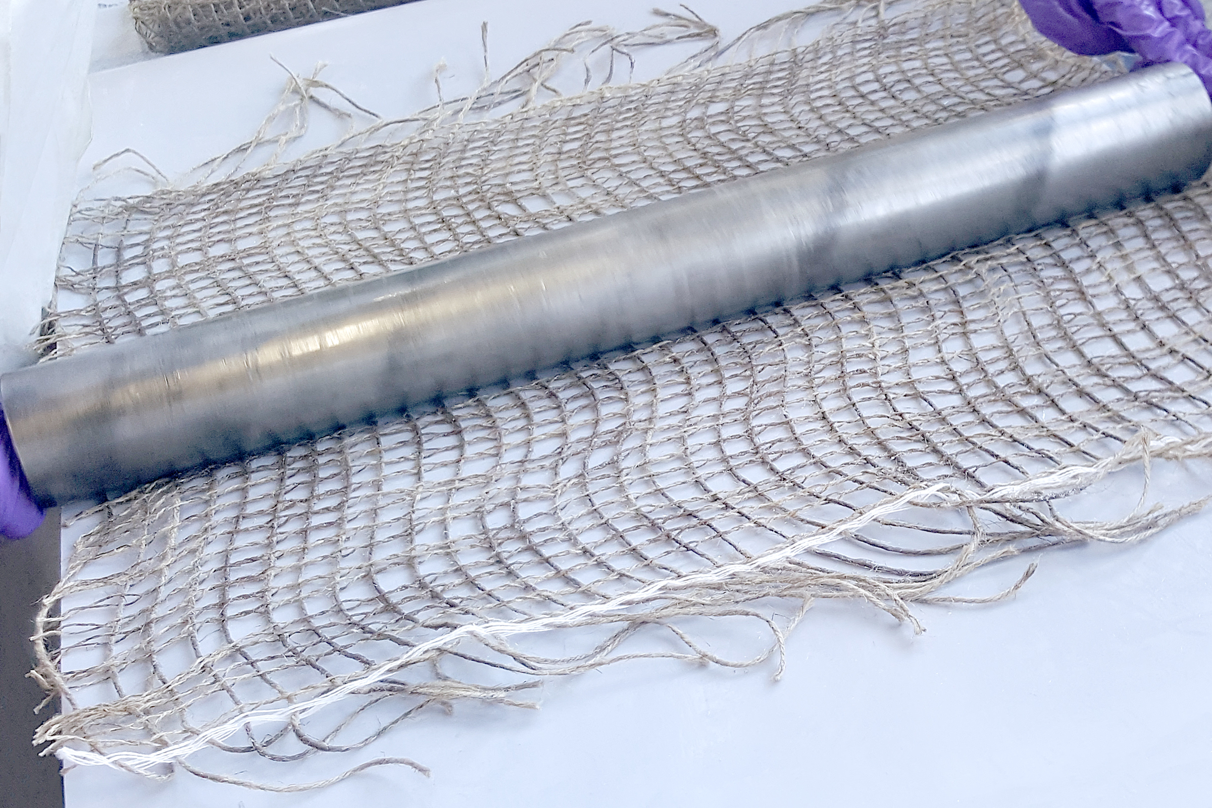 The photo shows a latticed flax fabric. A person is applying a coating to the fabric using a metal roller. 