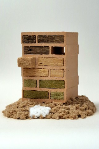 The photo shows a commercially available, vertically positioned masonry brick with 12 evenly spaced cavities. The cavities are filled with a variety of fiber-mat types. In front of the brick is a large pile of brown beech-wood fibers and a small pile of white bico fibers.