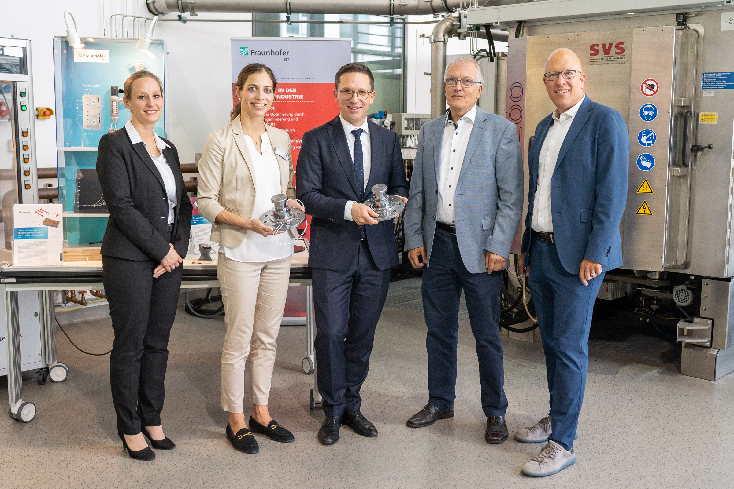 Science Minister Falko Mohrs visits the Fraunhofer IST The picture shows five people in the laboratory of the Fraunhofer IST. Two people are each holding a kingpin.