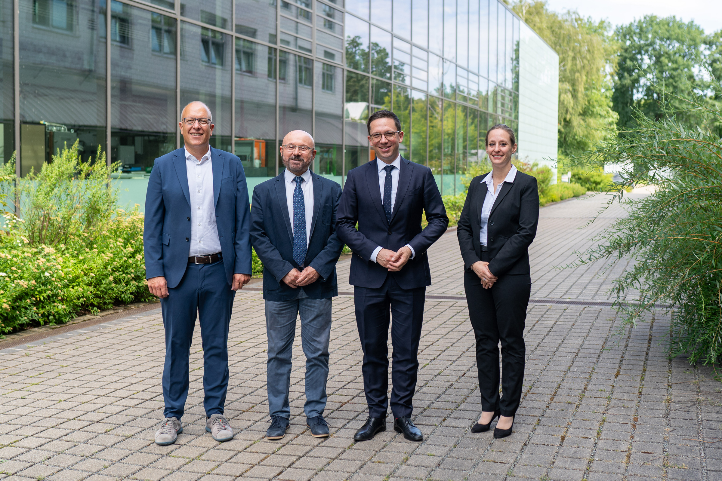 Science Minister Falko Mohrs visits the Fraunhofer campus in Braunschweig The photo shows four people on the outdoor area of the Fraunhofer Campus Braunschweig. A laboratory building with a glass front can be seen in the background.