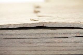 The photo shows the cut edge of a wooden panel that consists of several layers of beech wood which are glued to one another. The bonding has begun to detach, causing the layers of wood to separate from each other. The top layer has bulged and is fractured.  
