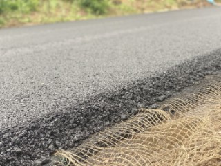 The photo shows a freshly laid layer of asphalt, under which lies a coarse-meshed fabric that protrudes from the edge of the asphalt layer and is therefore visible.