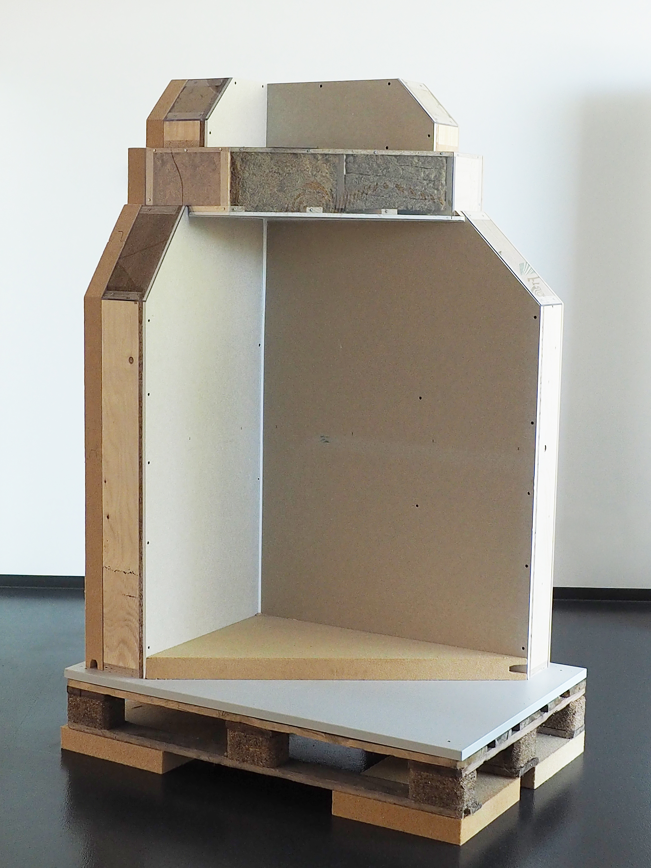 LIGNA 2025: Demonstrator for wall and floor construction made from renewable raw materials (“SusI4Chile” project) The photo shows a section of a building corner from the inside with a floor area of approximately 1 meter by 1 meter. The floor, walls, and ceiling are cut away in such a way that the insulation materials inside can be seen. All elements are made of various biomaterials, including wooden beams as the supporting structure, wood-based panels for cladding, and natural fiber insulation mats in the walls and floor.