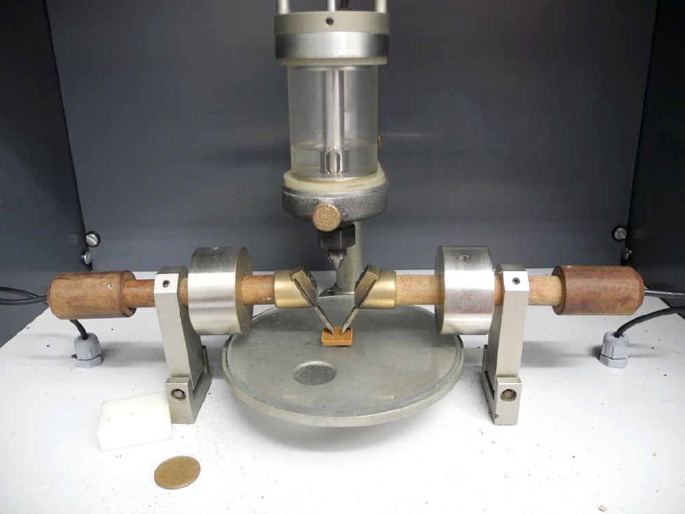 Testing apparatus with two electrodes touching a small test specimen made from bioplastic which is positioned in the center.  A drop-dispensing unit is suspended at a short distance above 