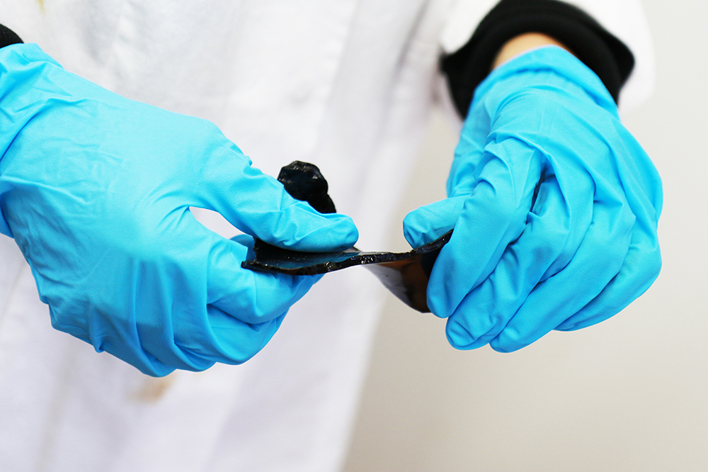 Two hands in blue laboratory gloves hold a black, plastic-like piece of lignin derivative and twist it in opposite directions.