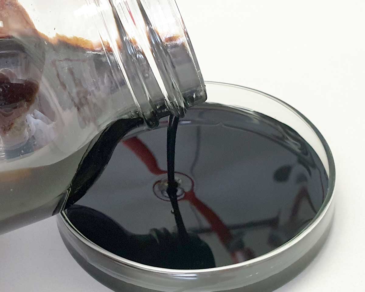 The photo shows a viscous, black-brown liquid being poured from a glass bottle into a laboratory dish.