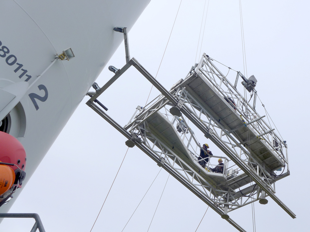 A large metal framework containing two people and technical equipment is pulled up the tower of a wind turbine using cables. 