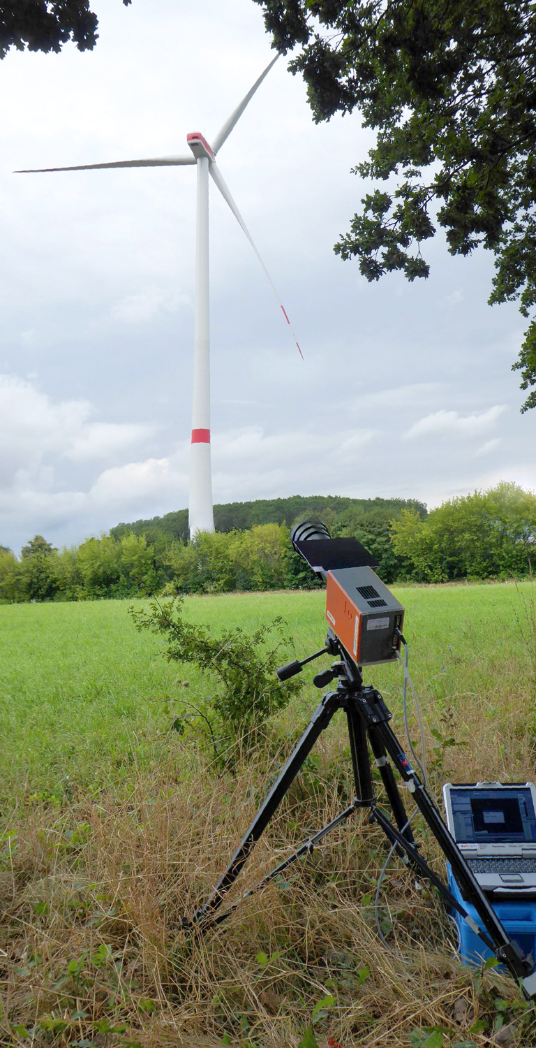 A camera on a tripod stands at the edge of a field. It is directed at a wind turbine which is around 100 meters away.