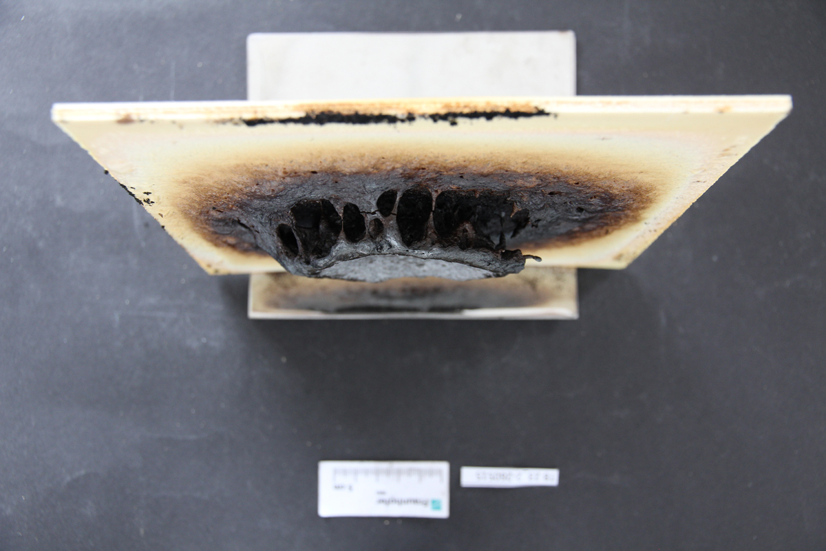 A piece of wood-based panel measuring 20 x 20 cm, from which a circular, foam-like structure rises in the center, which is heavily charred and blackened.