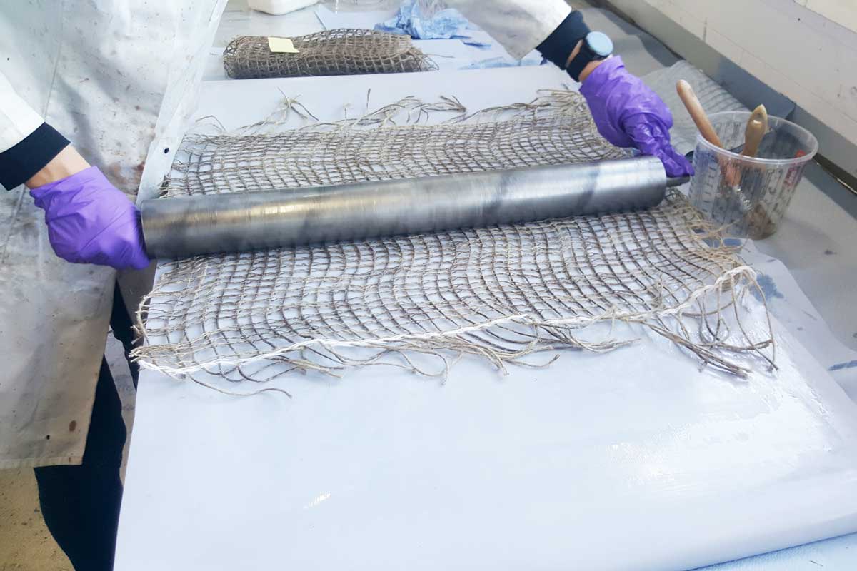 The photo shows a coarsely woven, brownish fabric which is lying on a workbench and which is being processed by a person using a steel tool that resembles a rolling pin.