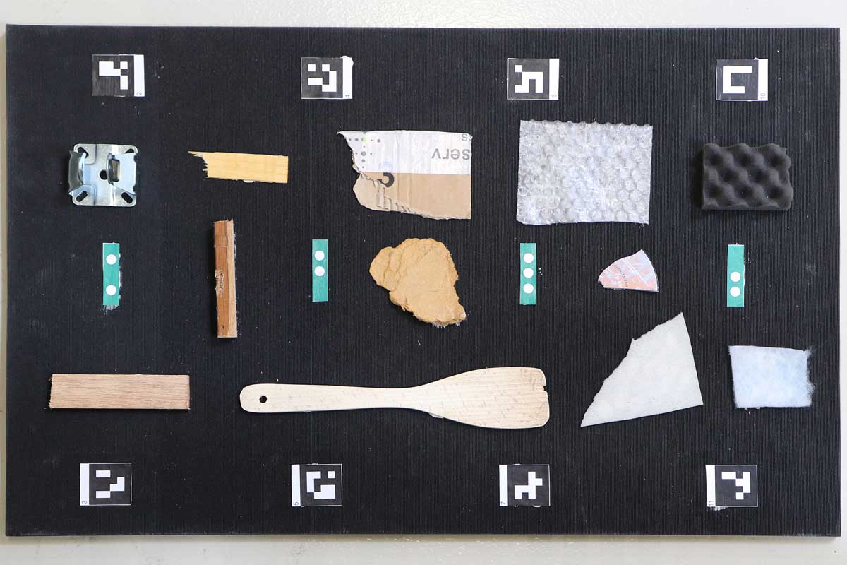  The photo shows a black rubber mat upon which diverse items of waste made from various materials such as wood, wood-based materials, plastic, cardboard and non-ferrous metal are laid out with a small amount of space around each item. On the upper and lower edges of the rubber mat are small pieces of paper with graphic, coarsely pixelated characters in black and white (ArUco markers). In the center of the rubber mat are small blue strips of paper, each with either two or three white dots.
