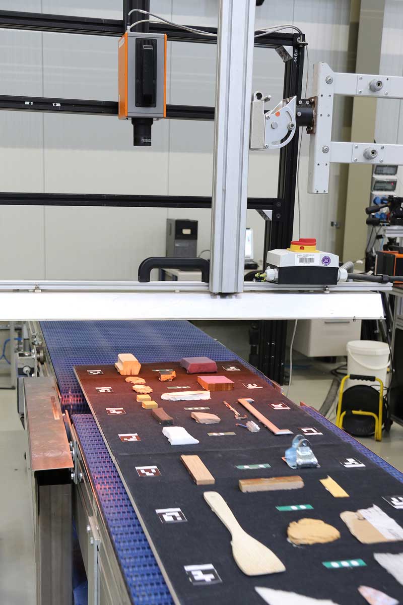  The photo shows various bulky waste samples laid out, together with graphic markers, on a black rubber mat which lies on a conveyor belt. Above the conveyor belt, a thermographic camera has been set up, which is directed vertically downwards onto the conveyor belt. 