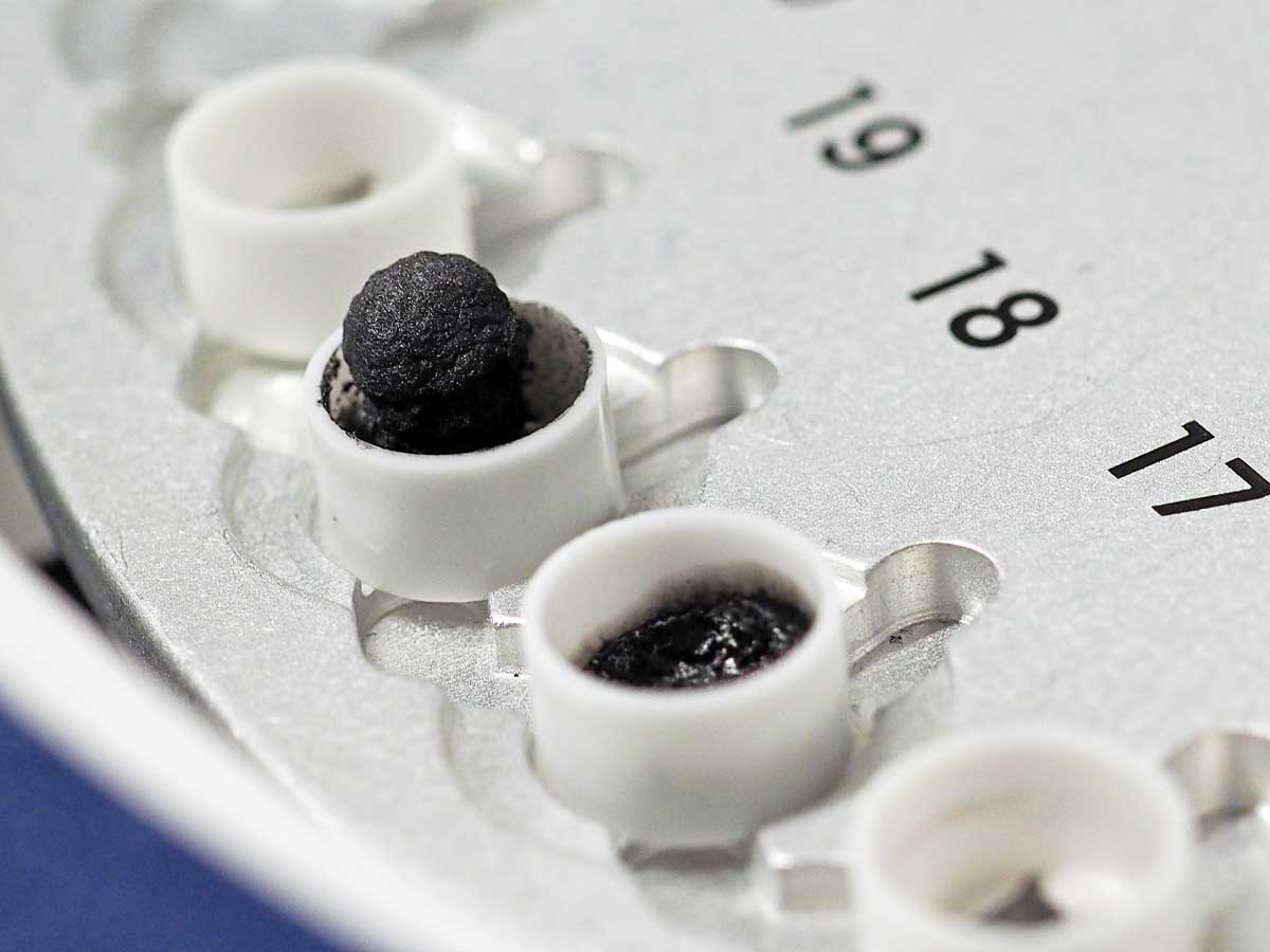 The photo shows several small, numbered crucibles in a measuring device. The crucibles contain varying amounts of a black, foam-like material.
