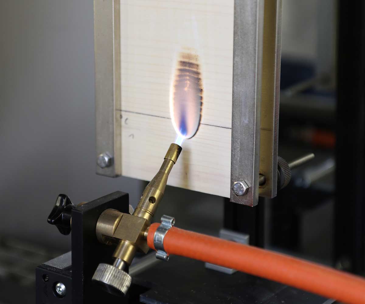 The photo shows a metallic apparatus in which a piece of spruce plywood is clamped. The piece of wood is being directly flamed by means of a gas flame which is positioned in front of it. On the wood, an oval dark brown patch (charring) is forming in the area of the flame.