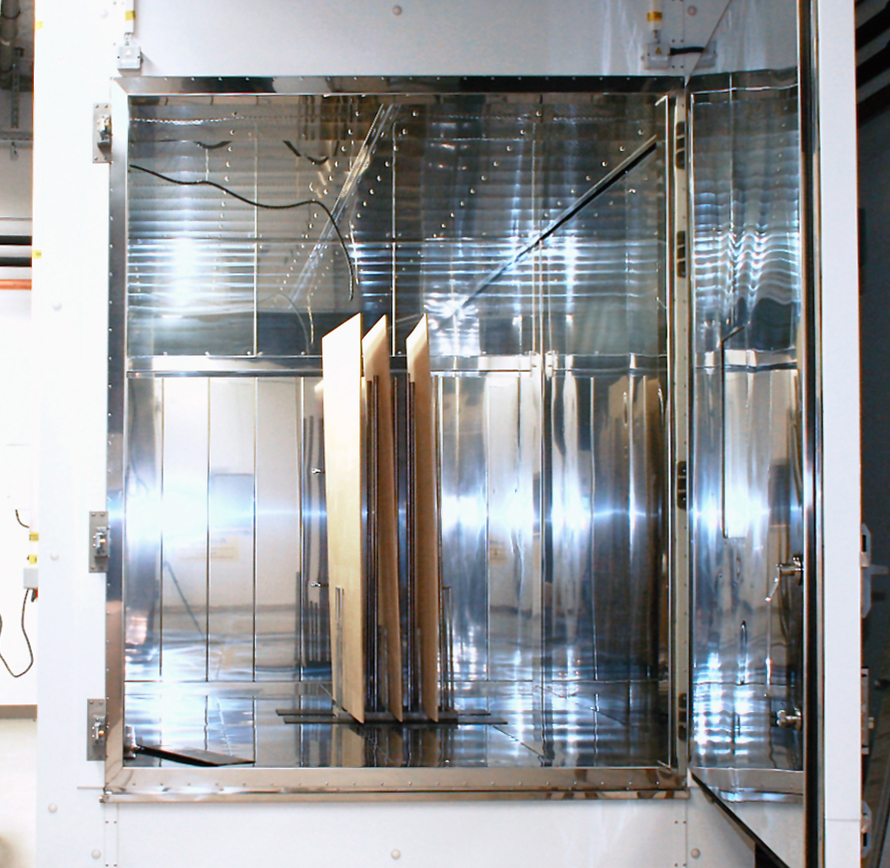 The photo shows a chamber that is approximately 3 meters wide, 4 meters deep and 3 meters high, lined on the inside with stainless steel. Through the open chamber door, a framework can be seen in the interior with three vertically positioned wood-based panels.