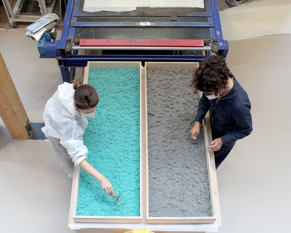 The photo shows two people in front of a press, each filling a rectangular flat mold with a colored wood-fiber mixture.