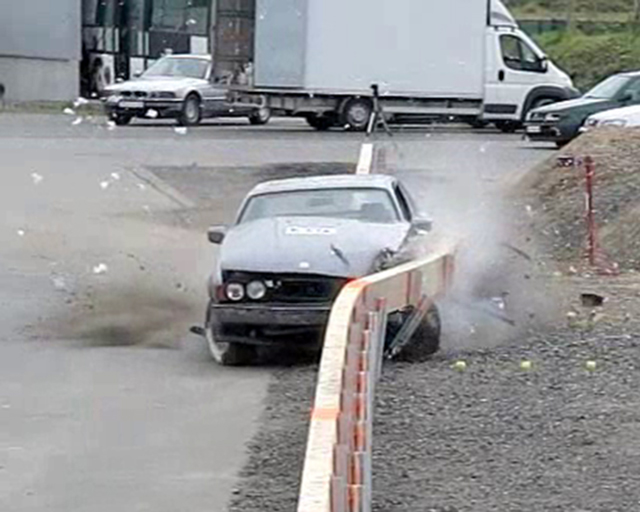 The photo shows a car from the front, as it crashes into a crash barrier made from wood. The crash barrier bulges out to the side by about 1 meter; a number of posts are torn out by the impact.