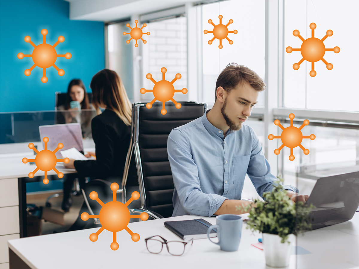 The photo shows an office with three computer workstations, each with one person sitting and working. Corona viruses are "floating" in the air (graphic visualization).