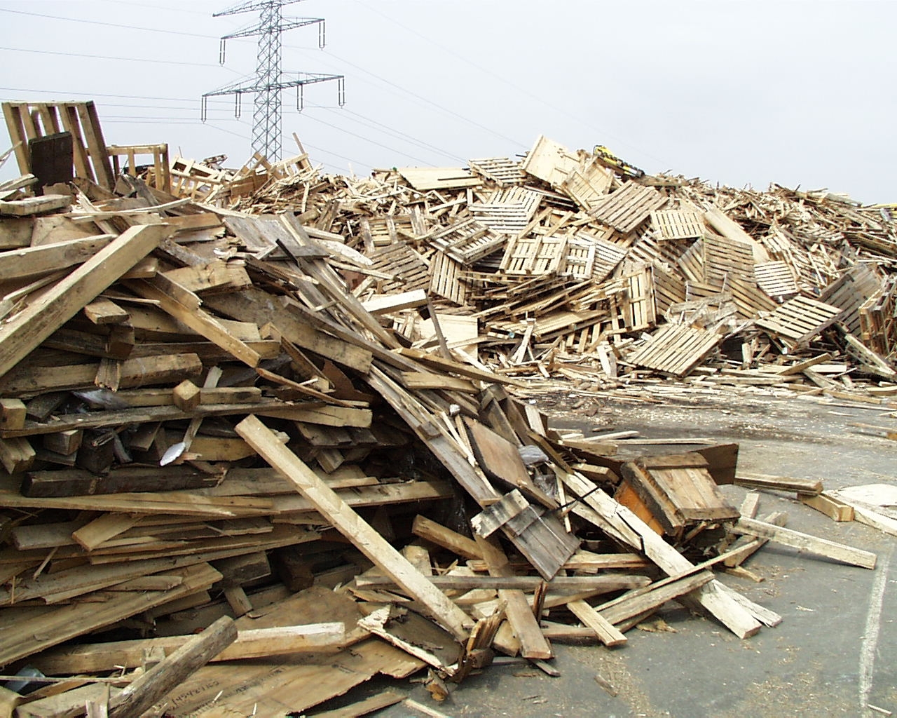 The photo shows large heaps of old wooden boards, wooden pallets and other wood products at a recycling yard.