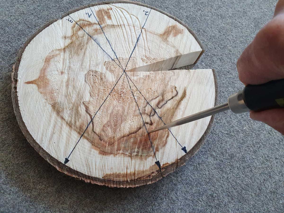 The photo shows a slice of spruce wood, partially darkened, lying flat. The surface is being depressed using a sharp hand tool.