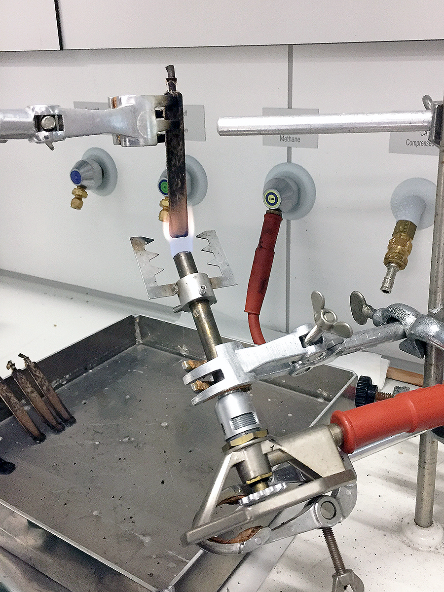 The photo shows an item of laboratory equipment: A WPC test rod is clamped vertically in a gripper arm and is being flamed from below at an angle by means of a gas burner.